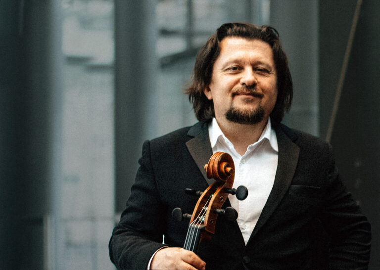 Der Cellist Friedrich Kleinhapl in einem Portraitfoto. Er trägt einen schwarzen Anzug und ein weißes Hemd ohne Krawatte und blickt Richtung Kamera. Er lächelt zufrieden und hält in der rechten Hand sein Cello.